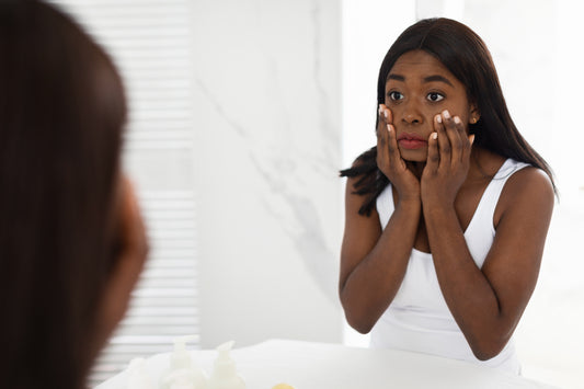 Confused Black Woman Looking At Her Dull Skin In Mirror And Touching Face