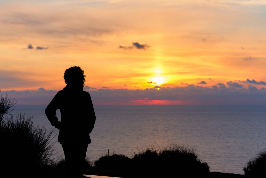 A person standing on a hill, gazing at the ocean during sunset. The image relates to Vitamin D Deficiency and Mental Health.