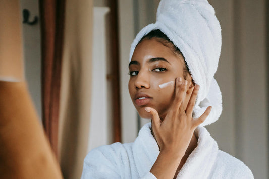 A woman in a towel gently applies cream to her face, using a gentle face cleanser.