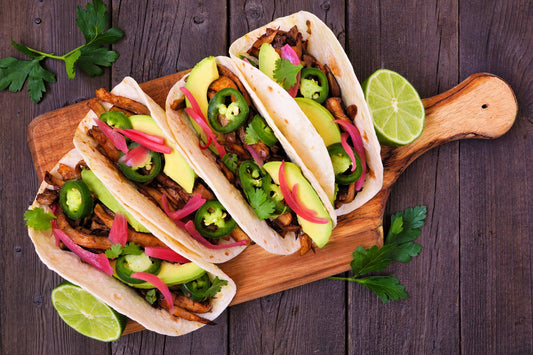 Three tacos made with plant-based ingredients, including mock meat, veggies, and lime, displayed on a wooden cutting board.