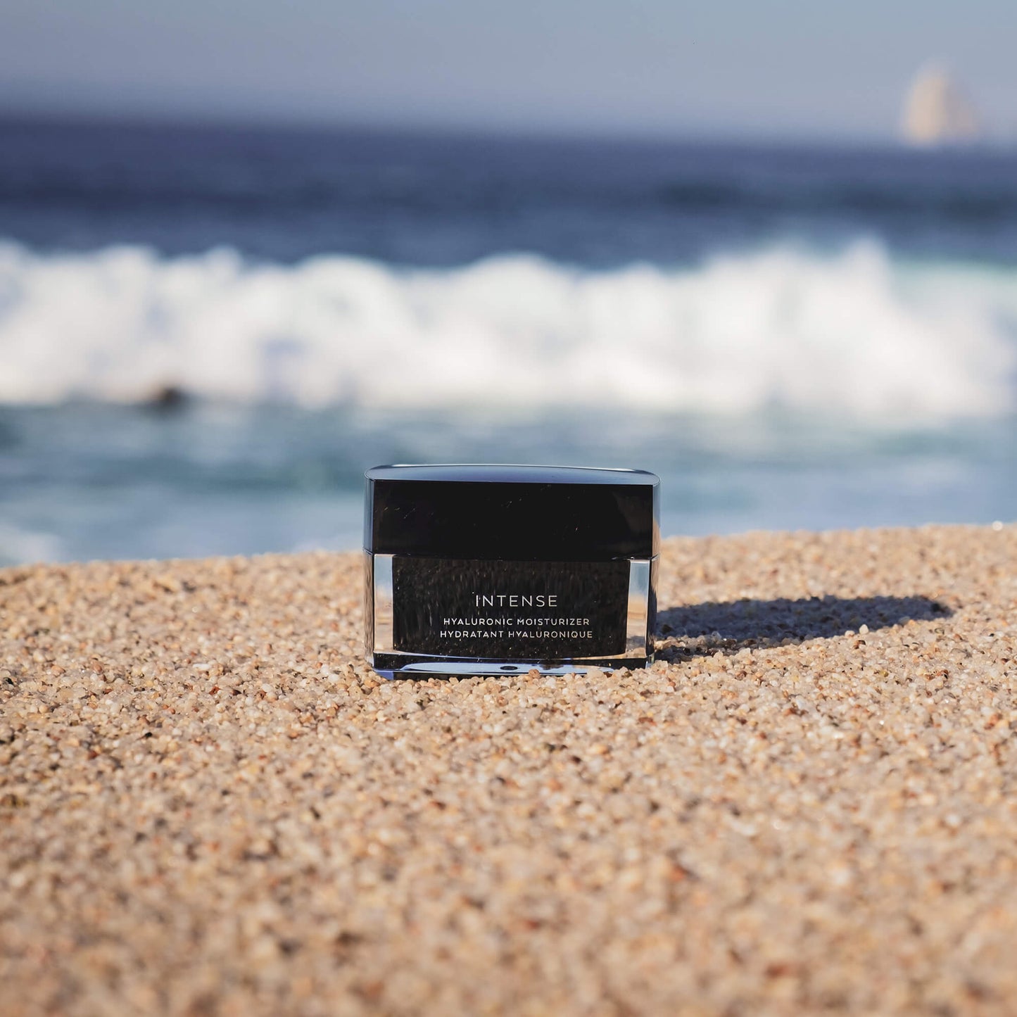 A jar of hyaluronic moisturizer, black in color, placed on sandy beach with the ocean in the background.